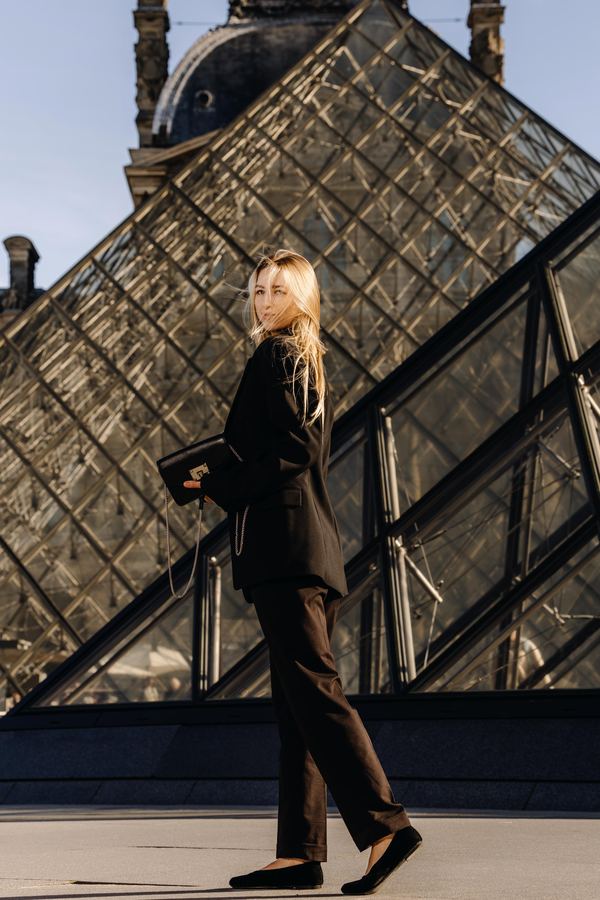 Portrait of a woman in black blazer and trousers, flat shoes, clutch, Louvre pyramid, Paris